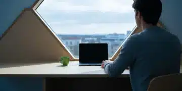 A person seen from behind, sitting at a modern birch wood desk with a laptop and green mug, looking out a large window at an urban skyline, representing the evolving landscape of modern living and work shifts.